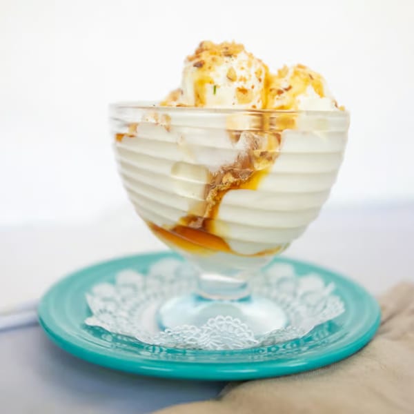 Vanilla ice cream with caramel drizzle and nuts in a glass bowl on a blue plate.