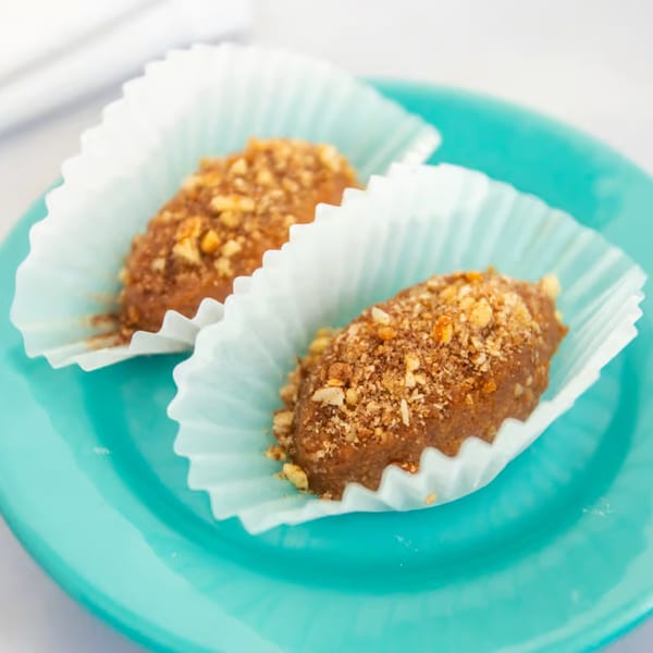 Two Greek melomakarona cookies, topped with nuts, in white paper cups on a turquoise plate.